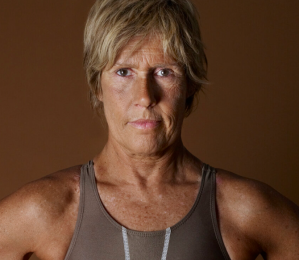 Diana Nyad with short hair wearing a sleeveless top stands against a brown background.